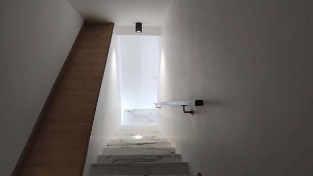 Eine Treppe mit Holzdielen führt zu einer Zwischenebene, an einer weißen Wand ist eine Lampe angebracht.