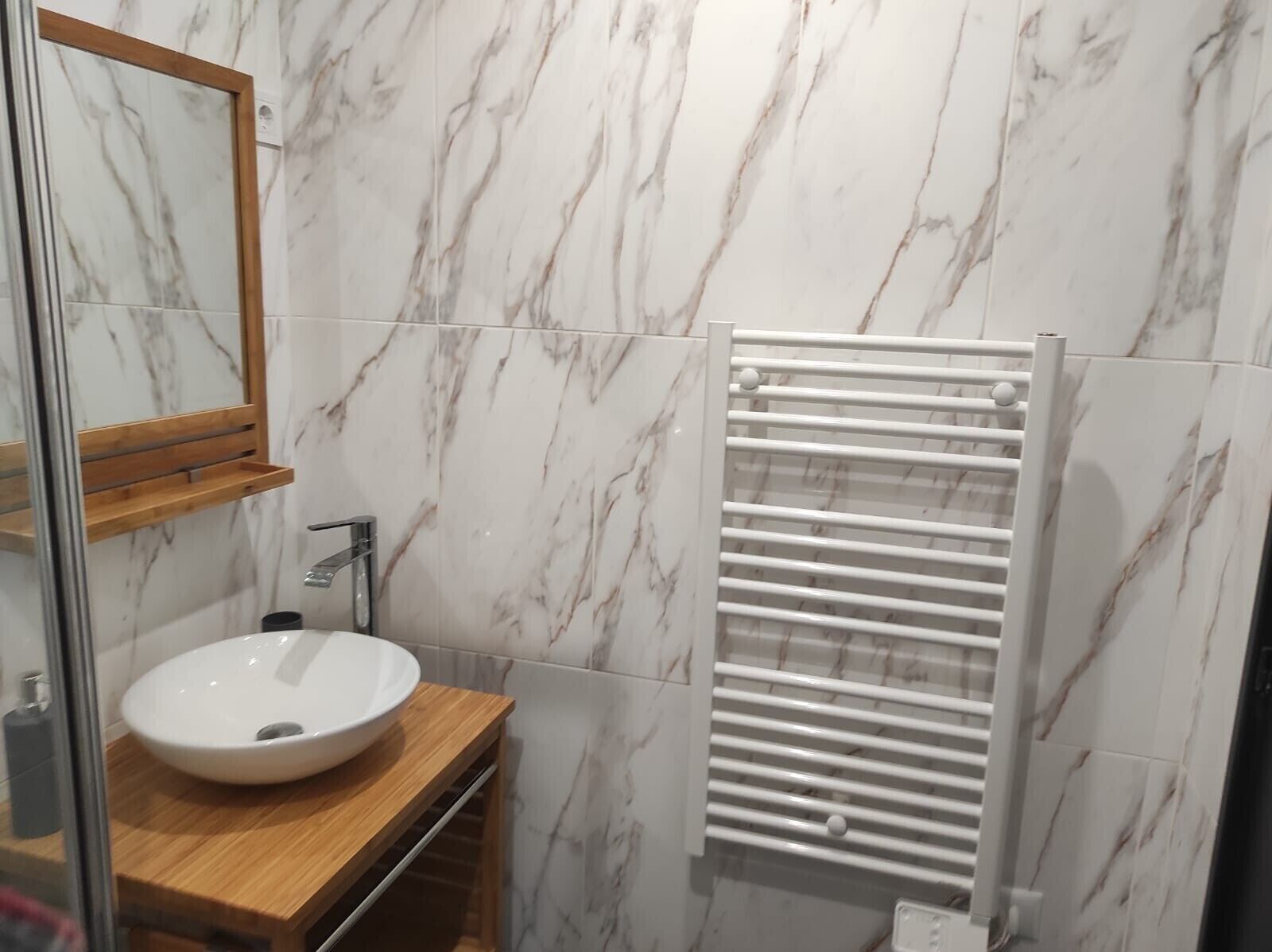 Un baño con pared de mármol, tocador de madera, lavamanos blanco, espejo y toallero blanco.