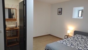 Ein Schlafzimmer mit Bett, Nachttisch mit Lampe, kleinem Wandspiegel sowie einem Badezimmer mit Waschbecken, Spiegel und Handtuchhalter.