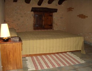 A bedroom with a bed, a wooden nightstand with a lamp, a window with wooden shutters, and a patterned rug. - Country House 'Casa Gimeno' with Mountain View, Wi-Fi and Air Conditioning (Bocairent)