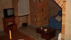 Quarto com lareira de tijolos, televisão sobre um suporte de madeira, uma cadeira azul e uma mesa pequena com uma vela.