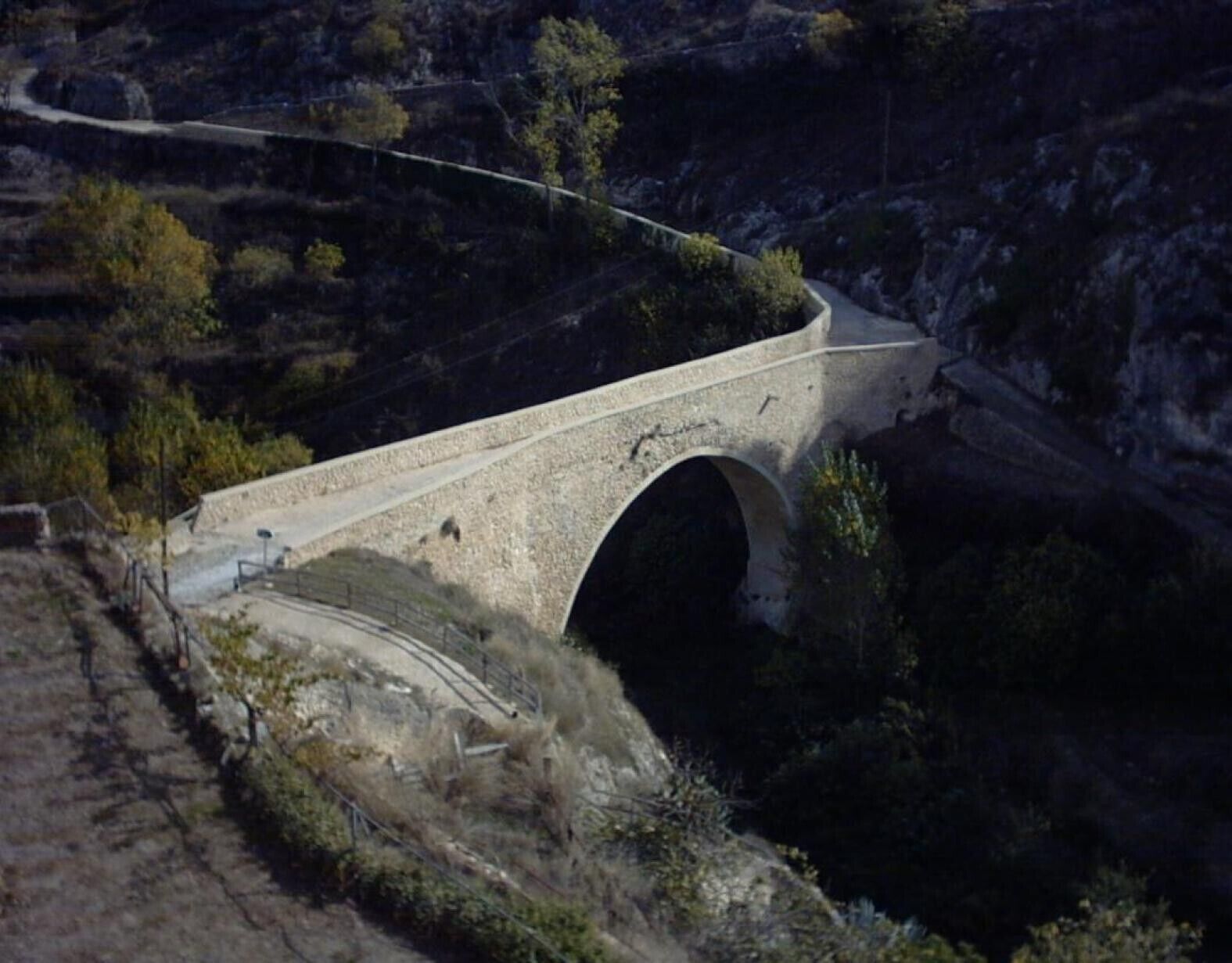 Un puente de piedra con arco sobre un valle, rodeado de árboles y terreno rocoso.