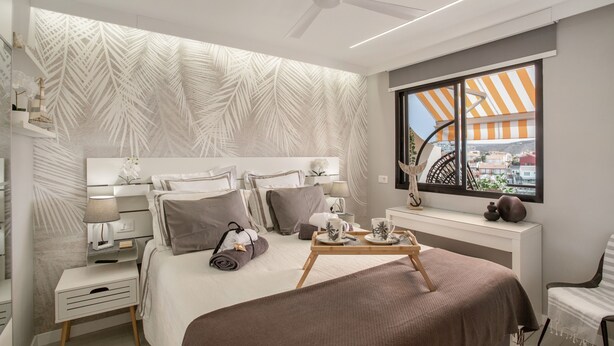 A modern hotel room with a bed, bedside table, lamp, and a view of the city through a window.