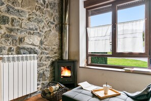 A stone wall, a wood-burning stove, a window with curtains, a cushioned bench, and a tray with a cup.