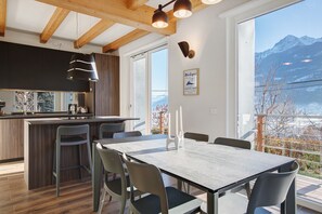 A modern kitchen with wooden beams, a dining area with a table and chairs, and a view of mountains through large windows.