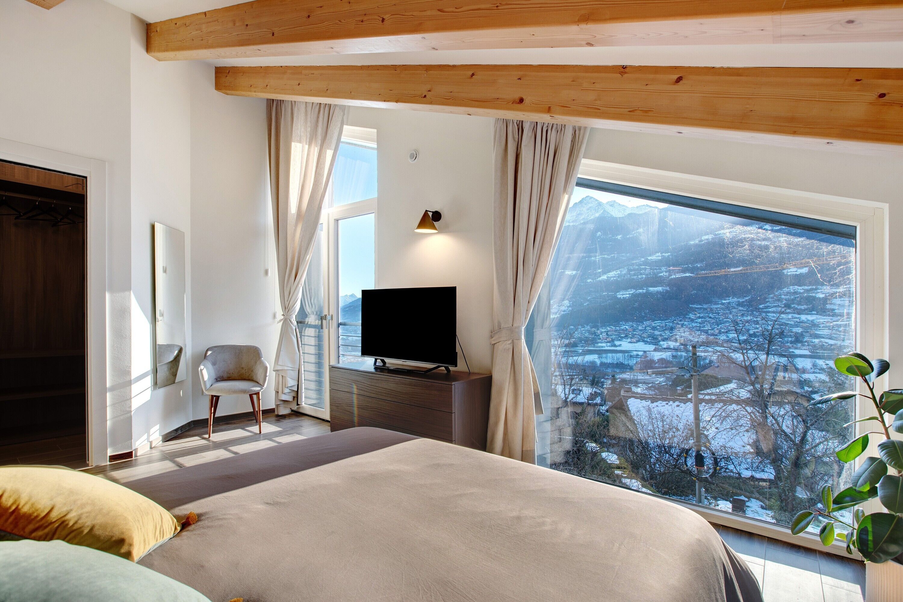 Een slaapkamer met een groot raam, een bed, een tv op een dressoir en een stoel.