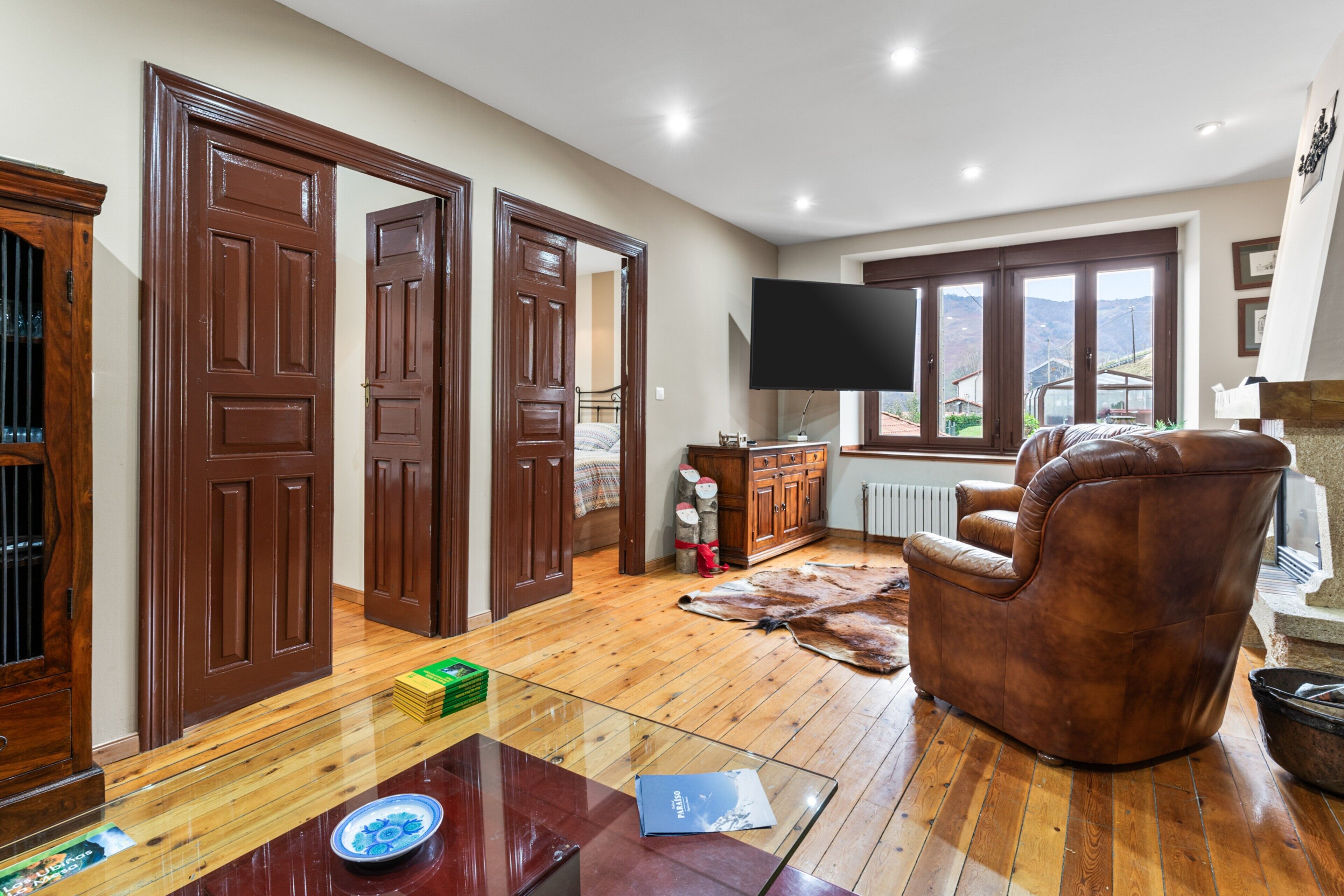 A spacious living room with wooden flooring, a leather armchair, a flat-screen TV, and large windows offering a view of the outdoors.