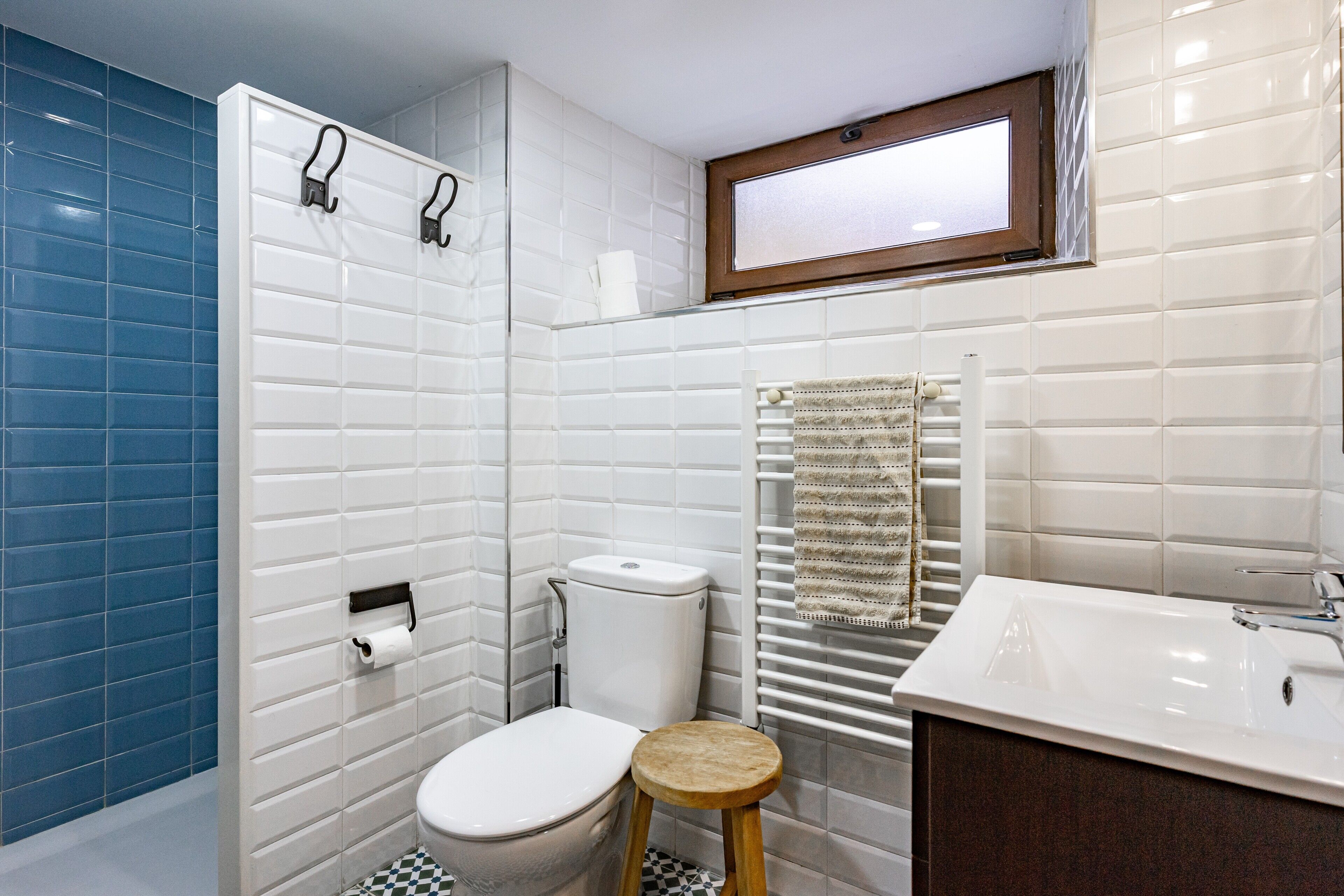 A bathroom with white tiled walls, a blue accent wall, a white toilet, a wooden stool, a white sink, and a towel rack.