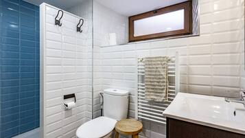 A bathroom with white tiled walls, a blue accent wall, a white toilet, a wooden stool, a white sink, and a towel rack.