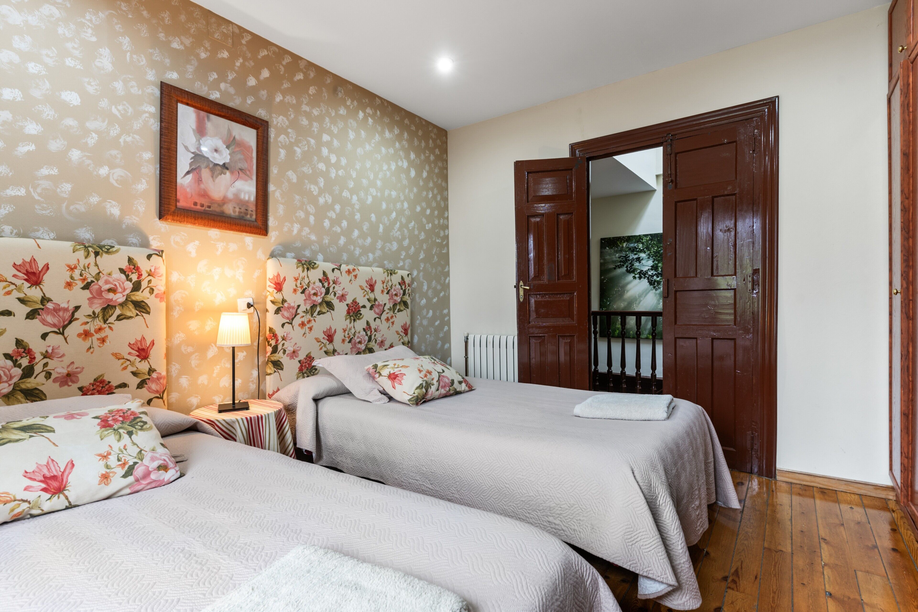 Une chambre avec deux lits, un papier peint à motifs floraux, un tableau encadré, une lampe et une porte en bois.