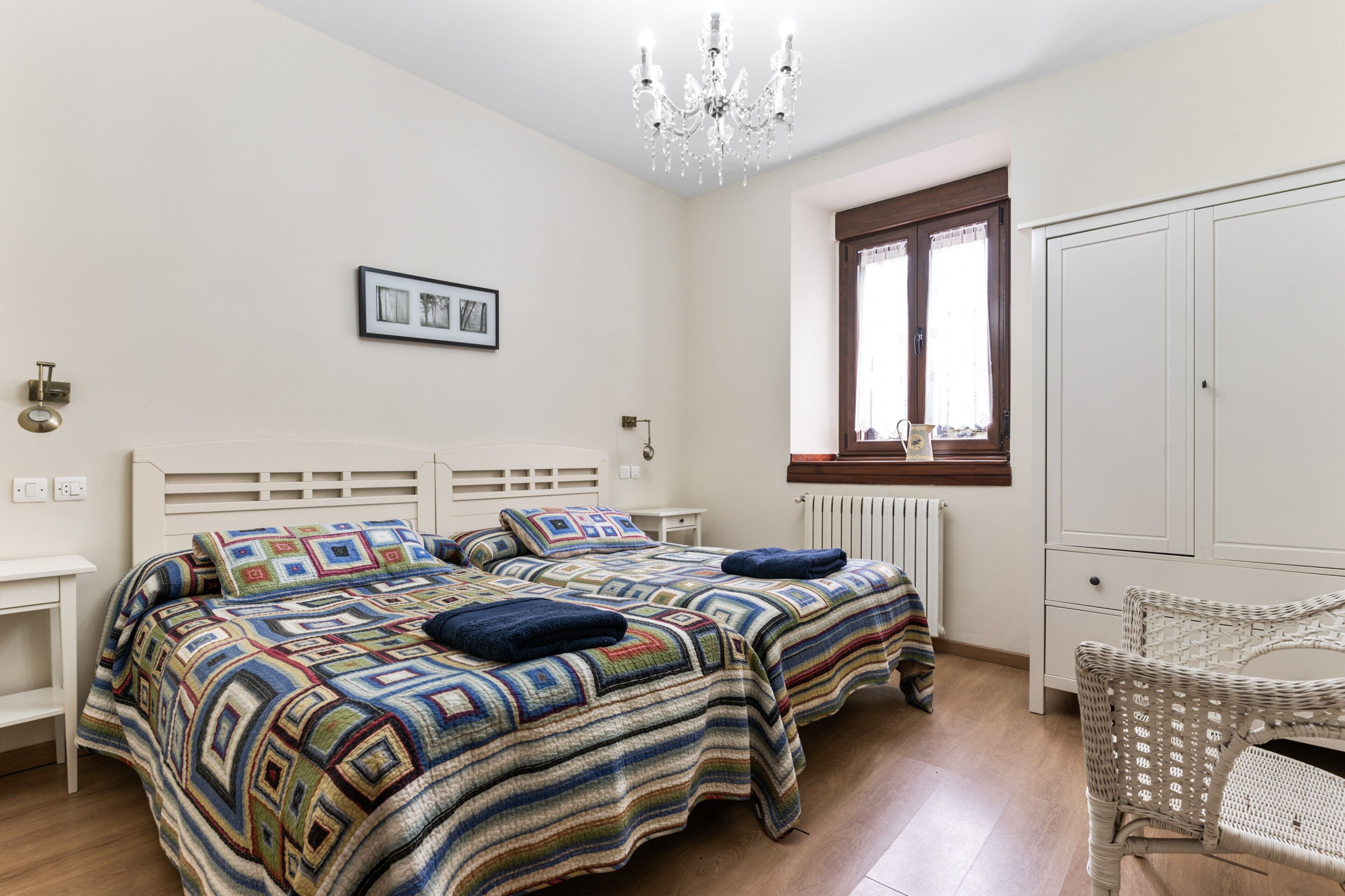 A bedroom with two beds, a chandelier, a radiator, a window, and a wardrobe.