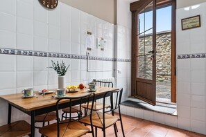 Une cuisine avec des murs recouverts de carreaux blancs, une table en bois avec des chaises, une fenêtre donnant sur un mur en pierre et une horloge au mur.