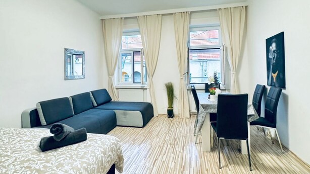 A modern room with a sectional sofa, a dining table with chairs, a potted plant, and a view of buildings outside. - Affordable 2BR Apartment; Close to Danube River (Vienna)
