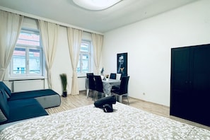 A bedroom with a bed, a sofa, a dining table with chairs, a potted plant, and a wardrobe.