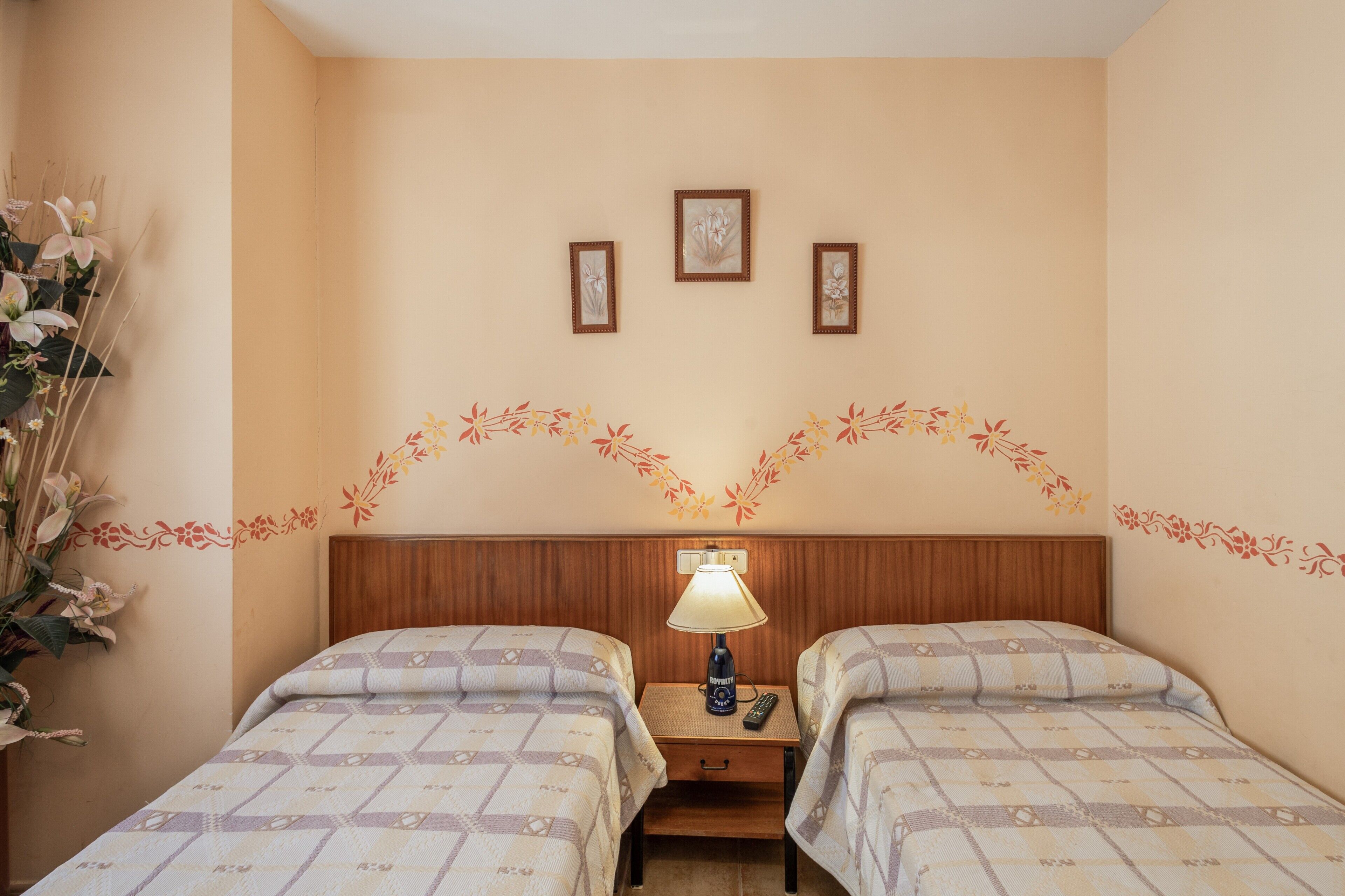 Habitación de hotel con dos camas, una mesita de noche con lámpara y tres cuadros enmarcados en la pared.