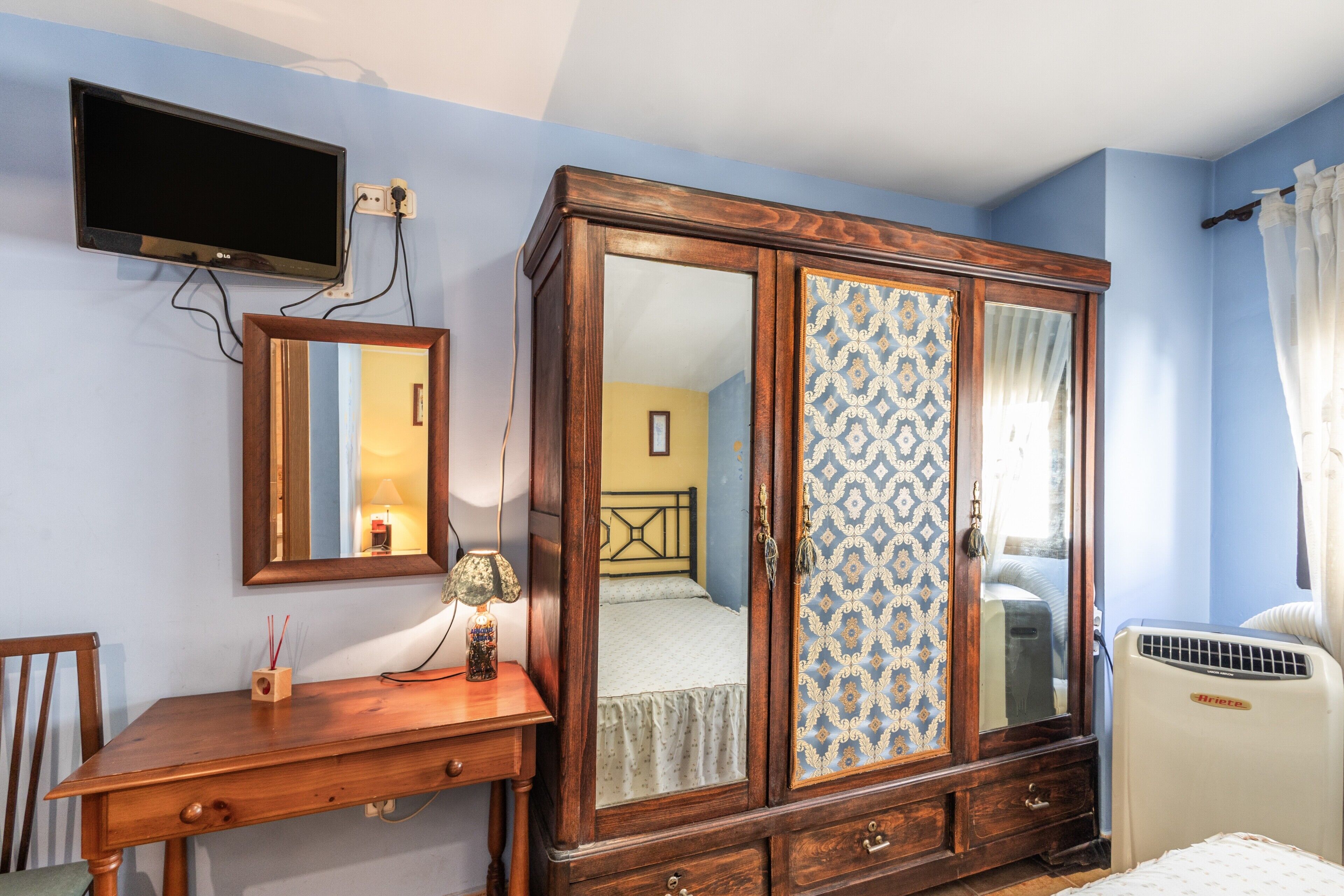 Un dormitorio con un armario de madera, una mesita de noche con lámpara, un televisor montado en la pared y una ventana con cortinas.