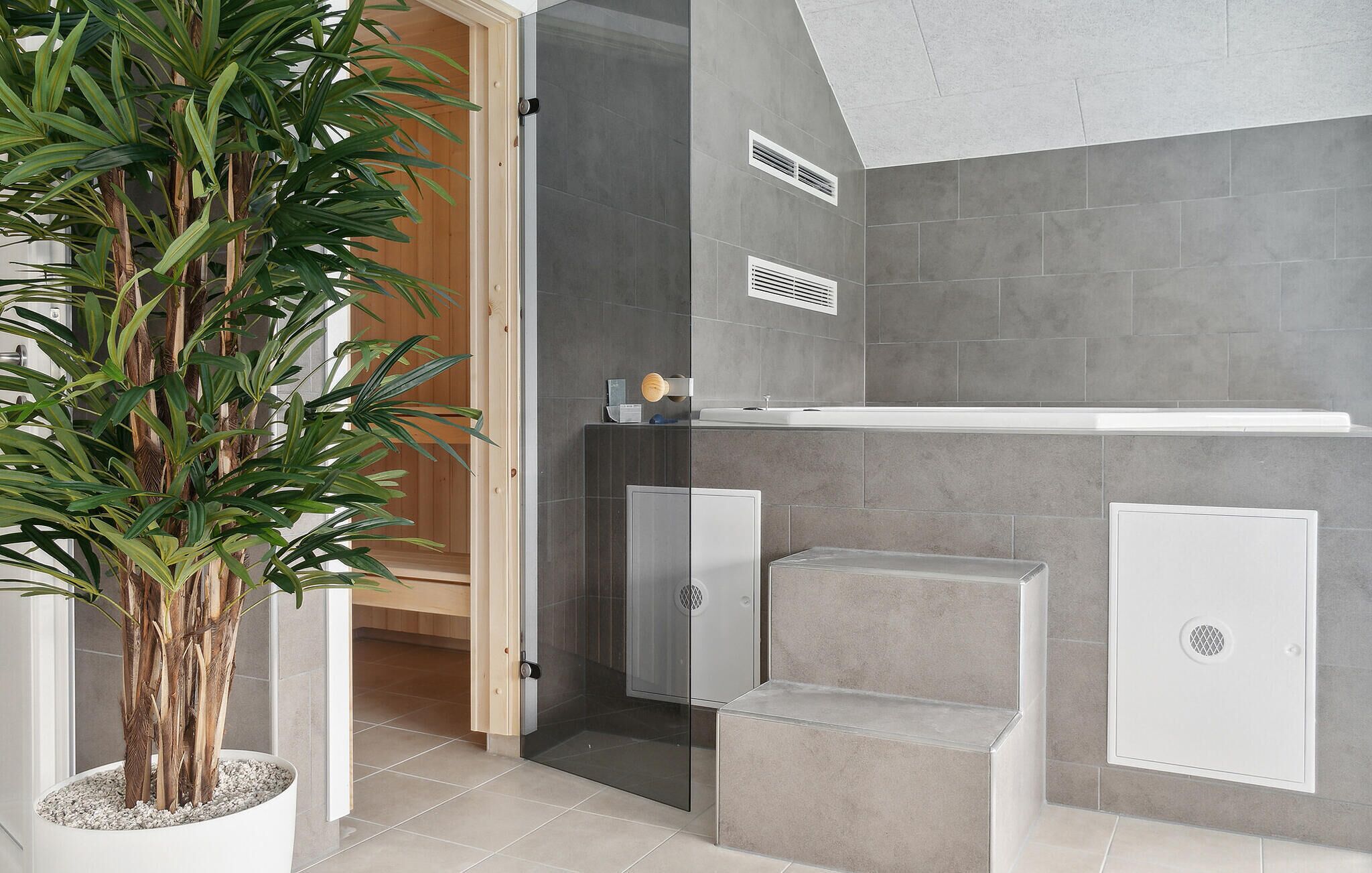 A modern bathroom with grey tiles, a glass shower enclosure, and a potted plant.