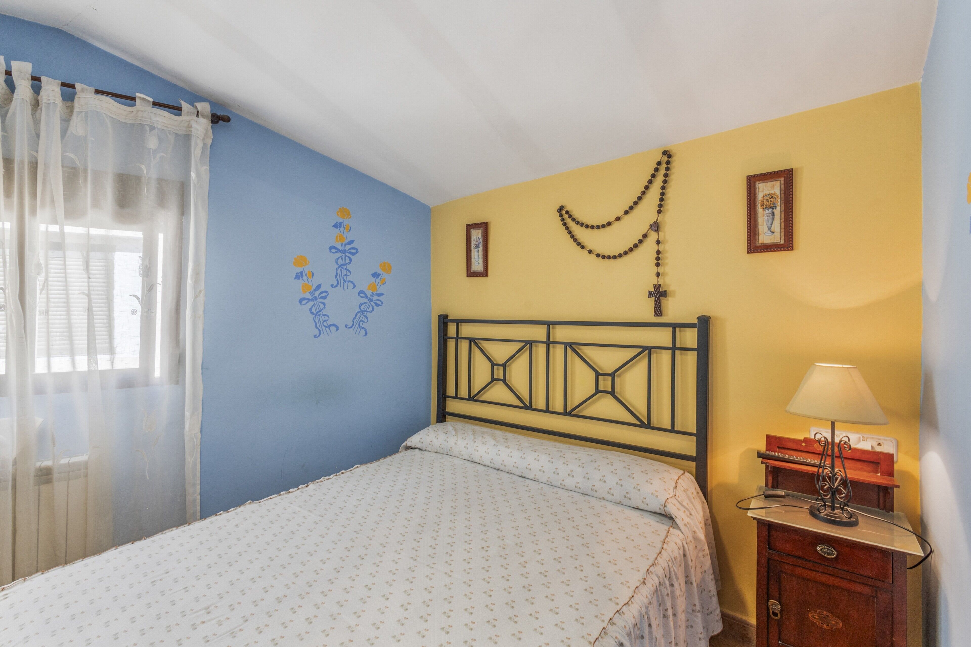 A bedroom with a bed, a nightstand, a lamp, a window with curtains, and wall decorations.