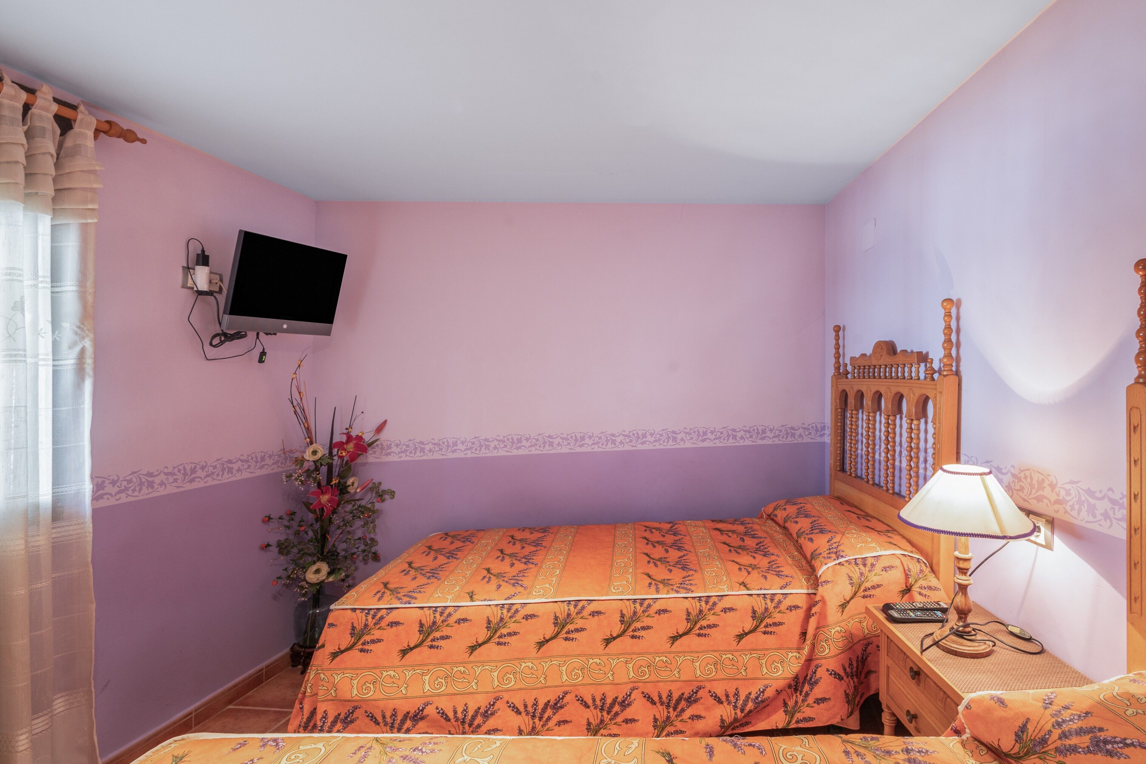 Une chambre à coucher avec un lit, une table de chevet avec une lampe, un téléviseur fixé au mur et un vase rempli de fleurs.