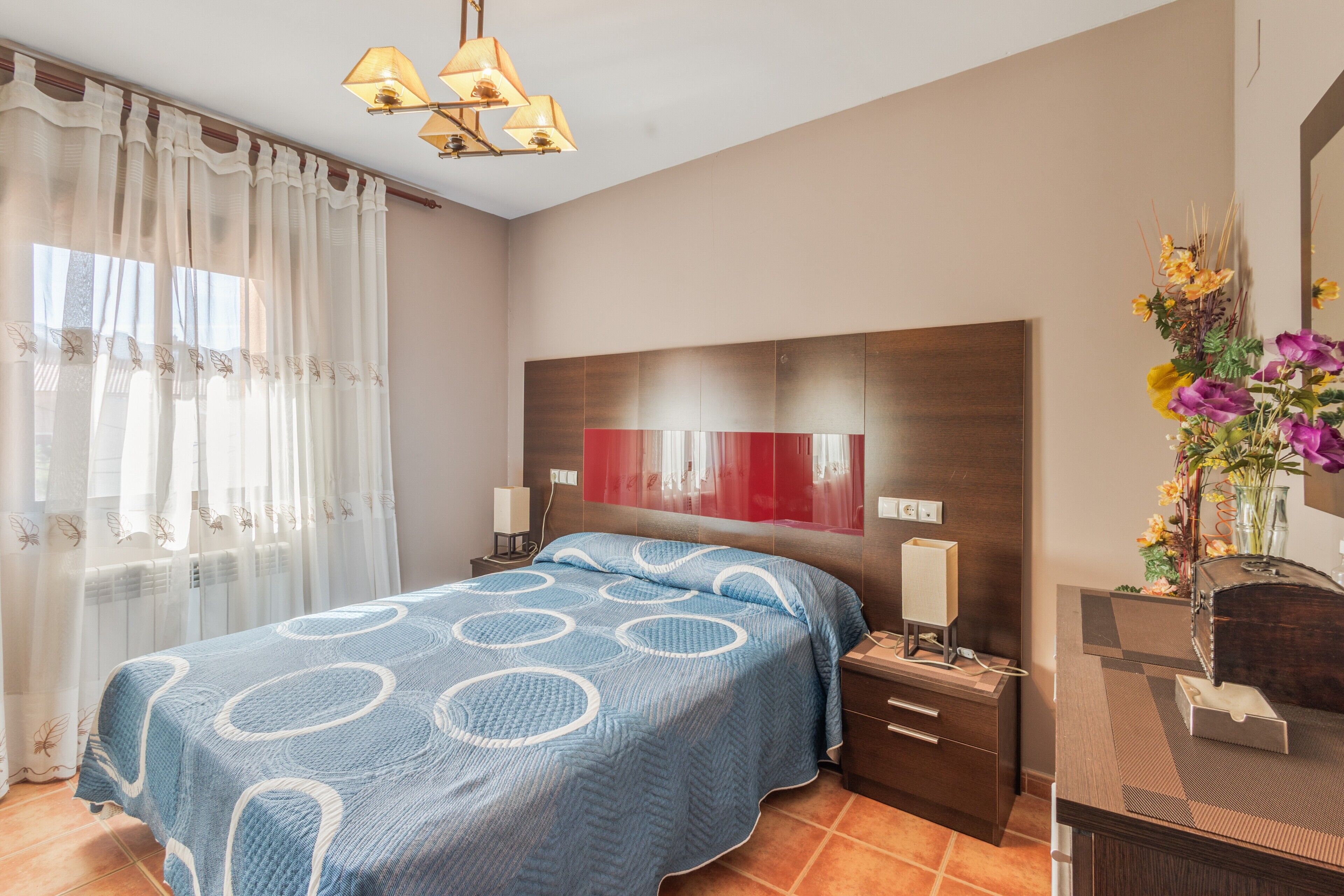 Un dormitorio con cama, mesitas de noche, un tocador, un espejo y una ventana con cortinas.