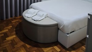 Un dormitorio moderno con una cama grande, una mesita de noche, un otoman redonda y un cuadro en la pared.