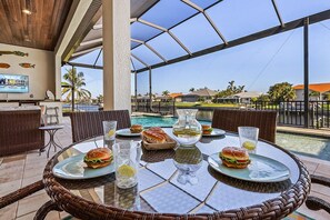 A dining area with wicker furniture, a glass table, and a view of a swimming pool and houses. - Stunning! Gulf Access, Heated Pool -Harbor Lights Hideaway - Roelens (Cape Coral)