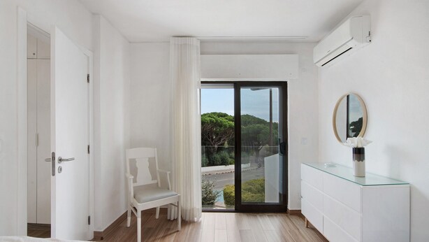 Quarto de hotel moderno com cama, poltrona, cômoda e porta de vidro deslizante que dá para uma varanda com vista para o verde.