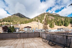 Un balcon doté d’un plancher en bois et d’un banc en métal, offrant une vue sur une ville avec ses bâtiments et une montagne.
