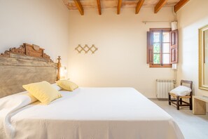 A bedroom with a wooden headboard, a bed with white bedding, a wooden chair, a radiator, and a window with wooden shutters. - Country House 'Can Segarut' with Private Pool, Private Terrace and Wi-Fi (Illes Balears)