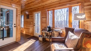 A cozy cabin interior with wooden walls, a leather sofa, a coffee table, and a large window offering a view of the outside.