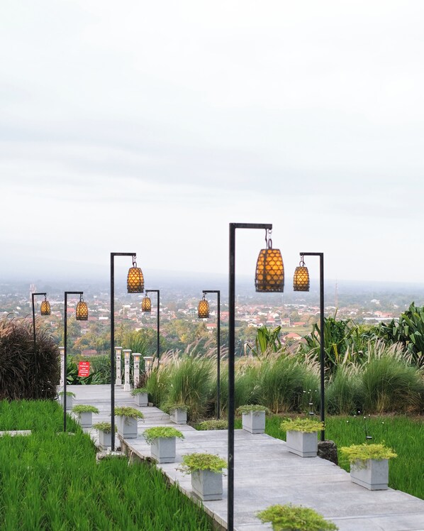 Un sendero flanqueado por follaje con faroles colgantes, con vistas a un paisaje urbano.