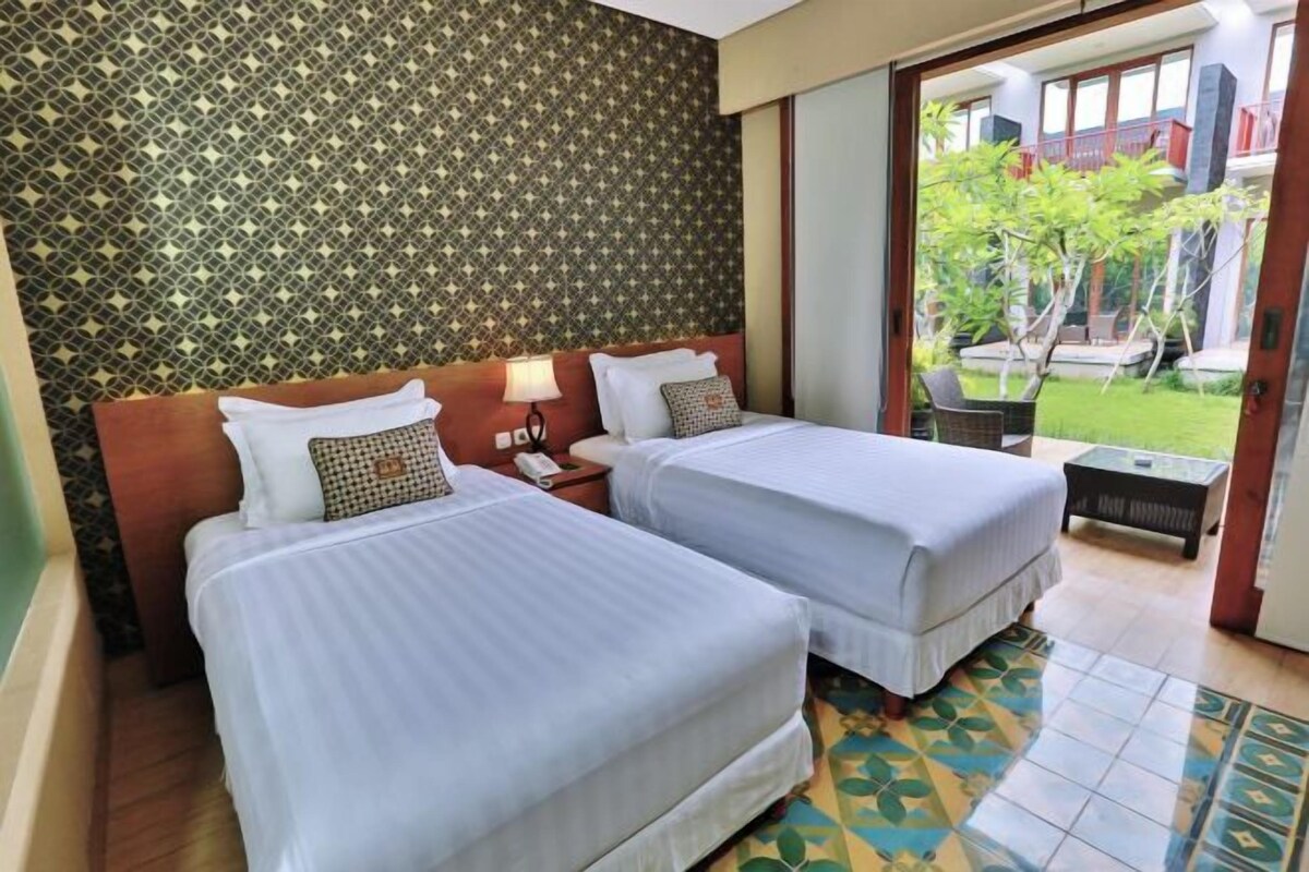 A hotel room with two beds, a patterned wall, a small table, and a view of a garden.