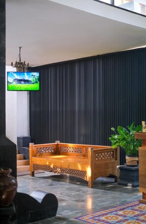 A room with a dark curtain wall, a wooden sofa with intricate carvings, a television mounted on the wall, and a potted plant.