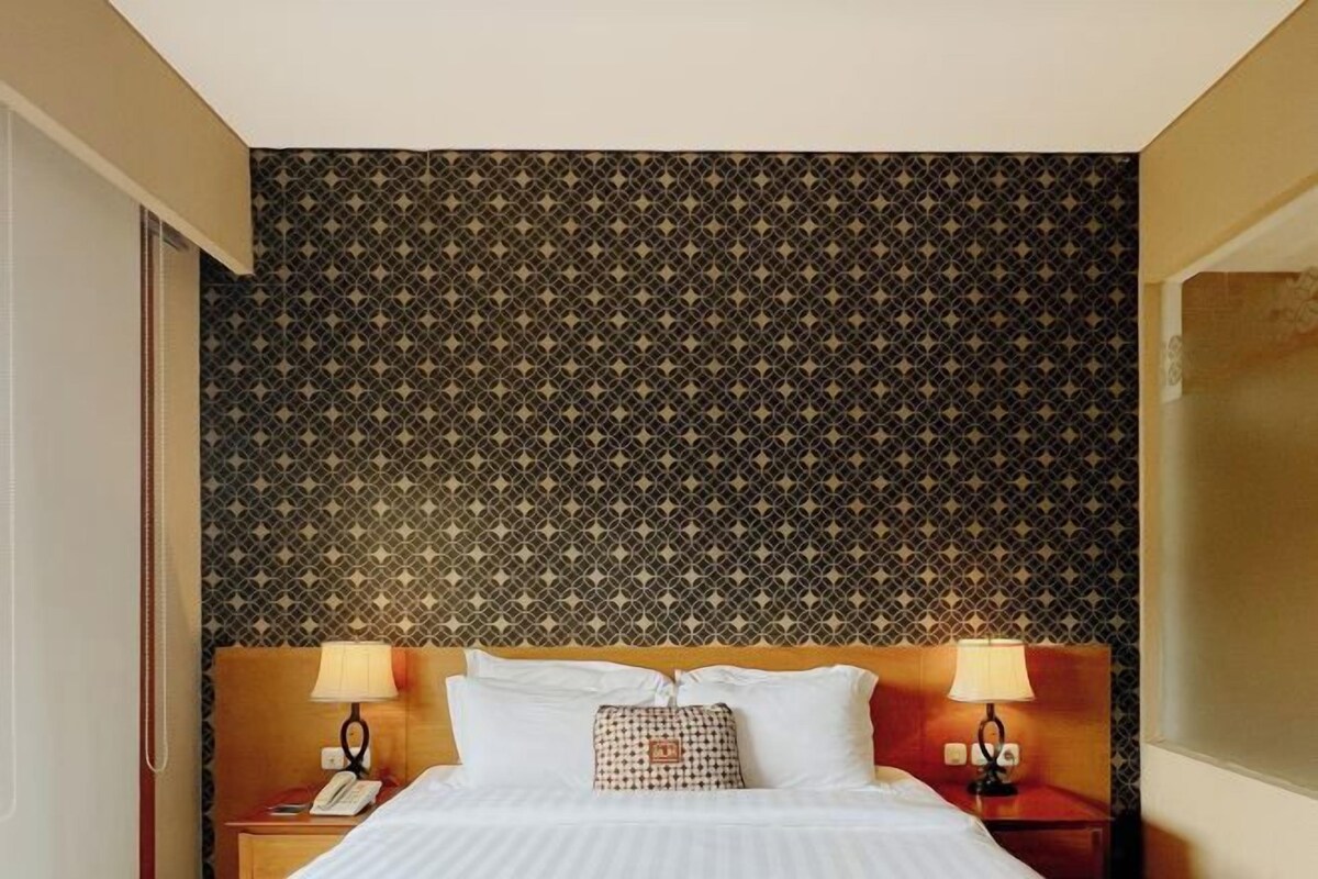 Una habitación de hotel con papel tapiz estampado, una cama con sábanas blancas, dos lámparas de noche y una mesita con un teléfono.