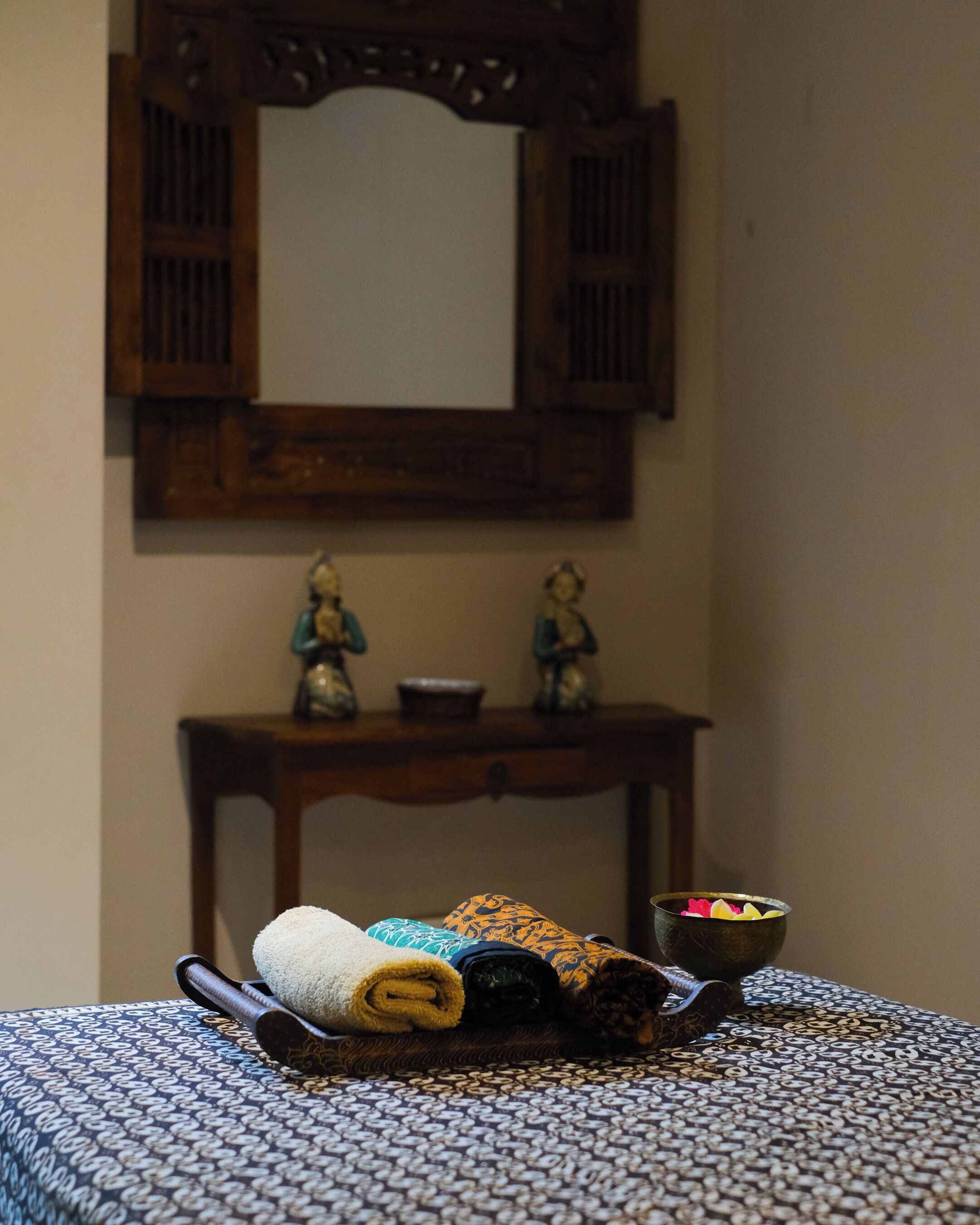 A bed with a patterned bedspread, a rolled-up towel, and a tray with a bowl and decorative items.