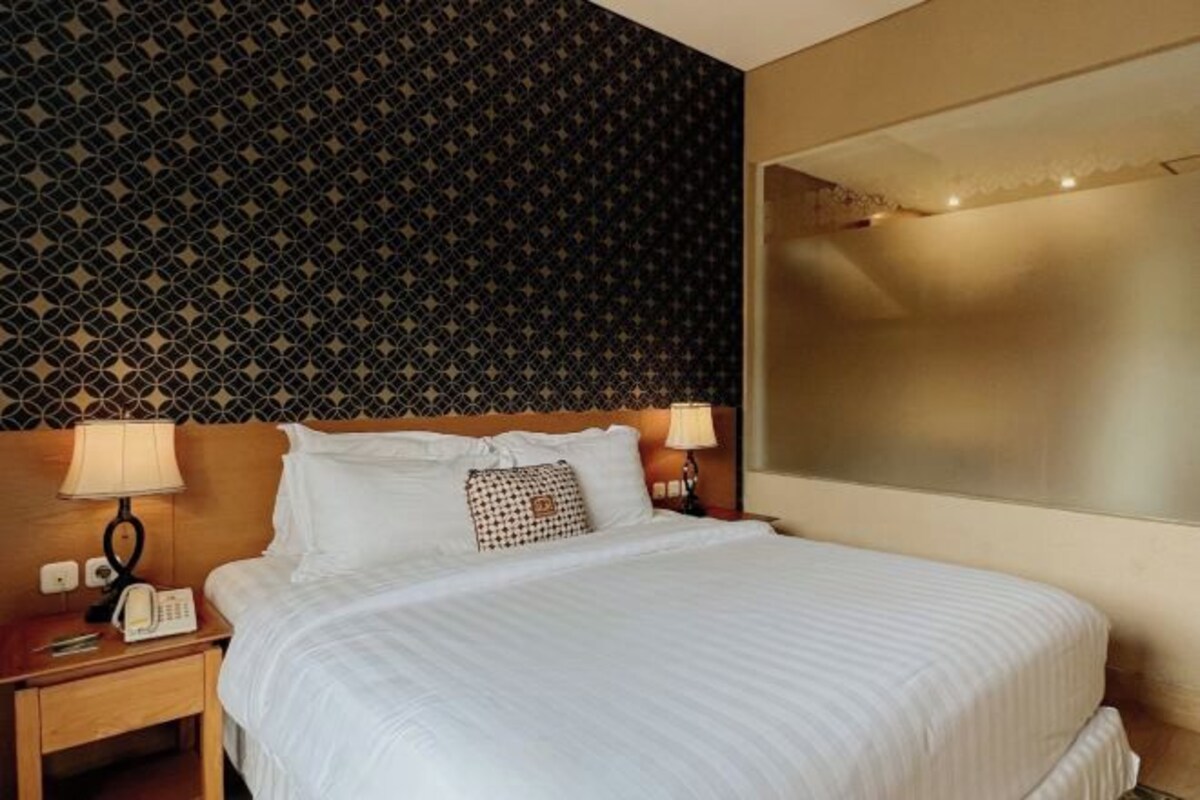 A hotel room with a patterned wallpaper, a bed with white linens, two bedside lamps, and a telephone on the nightstand.