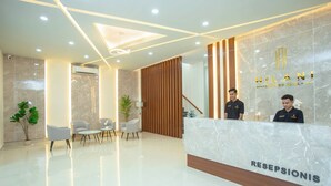 Ein moderner Hotel-Empfangsbereich mit einem Marmor-Empfangstresen, auf dem „HILANI BOUTIQUE HOTEL“ und „RECEPTIONIS“ stehen. Hinter dem Tresen befinden sich zwei Rezeptionisten, ein Raumteiler aus Holzleisten und eine Sitzecke mit Stühlen und einem Tisch.