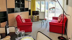 A compact living space with two red sofas, a dining table with chairs, a TV, and a large window with a view of greenery. - Cosy apartment for 3 people with WIFI, TV, balcony, pets allowed and panoramic view (Grindelwald)