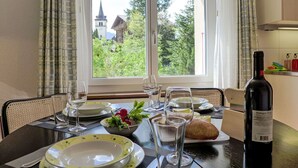 A dining table set with plates, glasses, and a bottle of wine, with a window view of a church and greenery. - Cosy apartment for 3 people with WIFI, TV, balcony, pets allowed and panoramic view (Grindelwald)