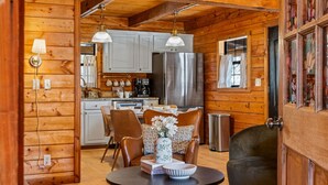 A cozy cabin interior with a wooden interior, a dining area with wicker chairs, a round table, and a kitchen with stainless steel appliances. - Heated Pool + Theater + Arcade | Near Dollywood (Sevierville)