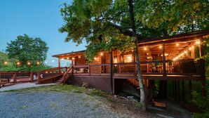 A wooden cabin with a covered deck and outdoor seating area, surrounded by trees. - Heated Pool + Theater + Arcade | Near Dollywood (Sevierville)