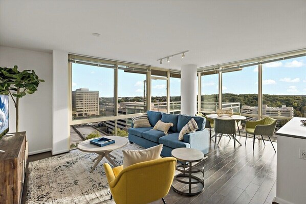 A modern living room with a blue sofa, yellow armchair, and round coffee table. Large windows offer a city view. - Tysons 2BR | Corner Views, Balcony, Pool & Gym (Tysons)