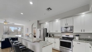 A modern kitchen with white cabinets, stainless steel appliances, a marble countertop, and a bar area with stools. - The Bridges? How Fancy! Waterview, Garage, Pool. (Chandler)