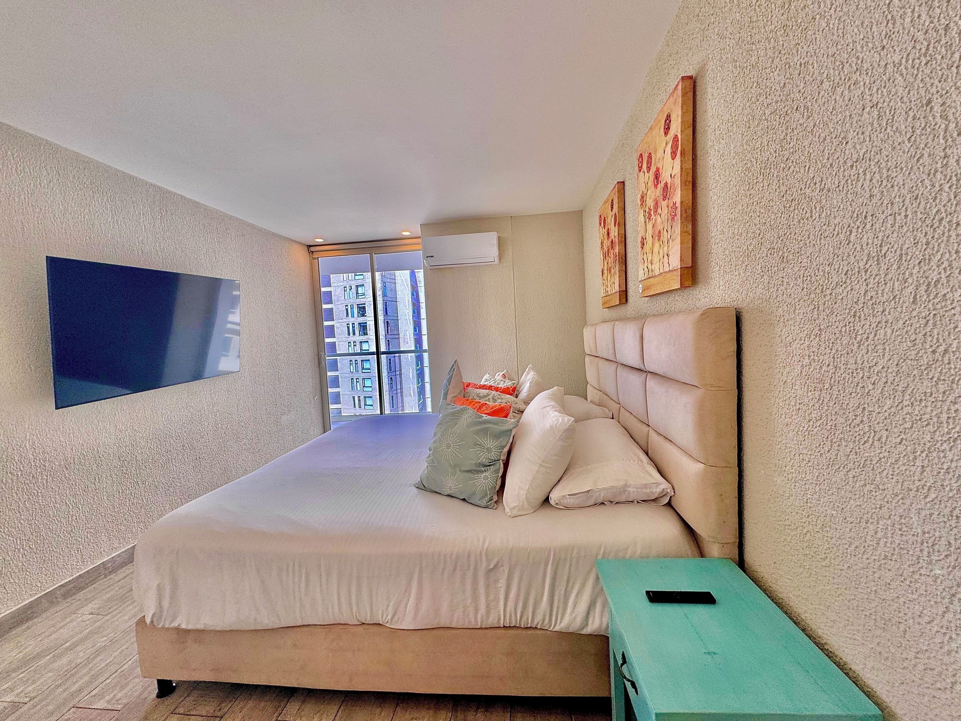 A modern bedroom with a large bed, a TV mounted on the wall, and a view of a building through the window.