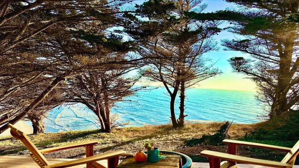 A wooden deck with two wooden chairs and a small table, overlooking a body of water with trees in the background.