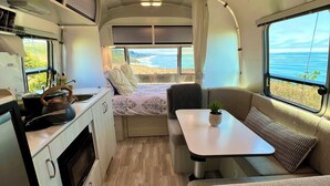 A compact RV interior with a kitchenette, a dining area, and a bed with a view of the ocean.