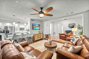 A spacious living room with leather sofas, a ceiling fan, a dining area with a kitchen in the background, and a large TV.
