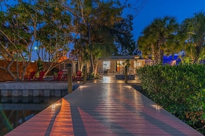 A wooden pathway leading to a building with a pool, surrounded by tropical vegetation.