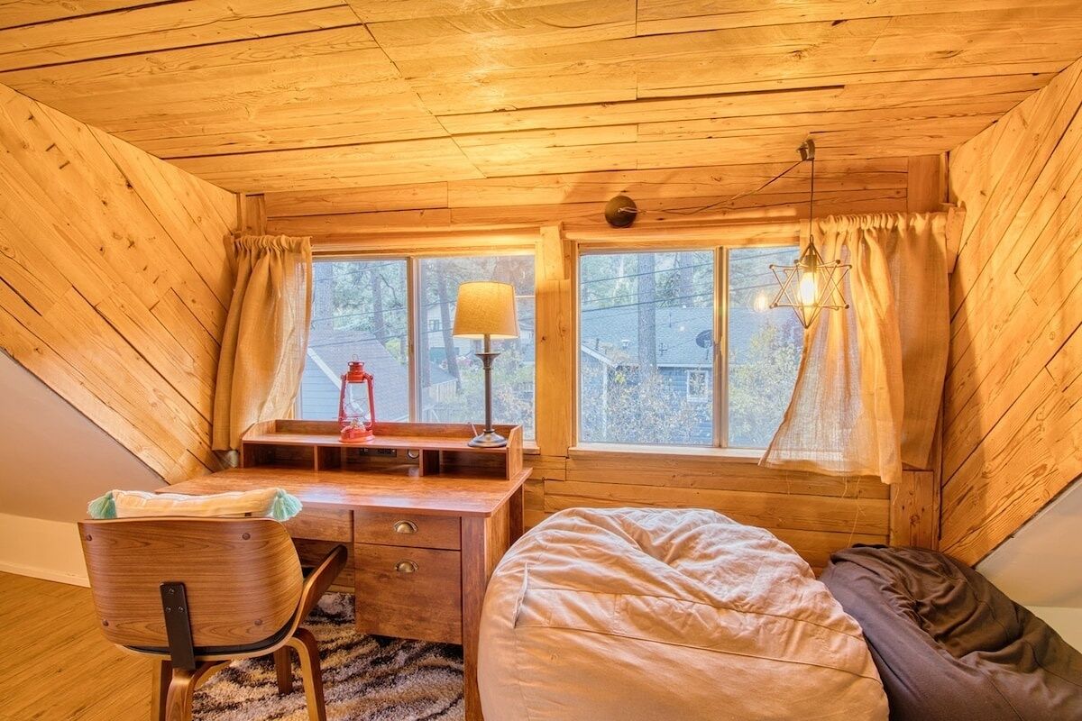 A cozy attic bedroom with a wooden sloped ceiling, a desk with a lamp, a bed, and two windows with curtains.