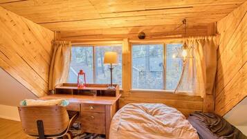 A cozy attic bedroom with a wooden sloped ceiling, a desk with a lamp, a bed, and two windows with curtains.